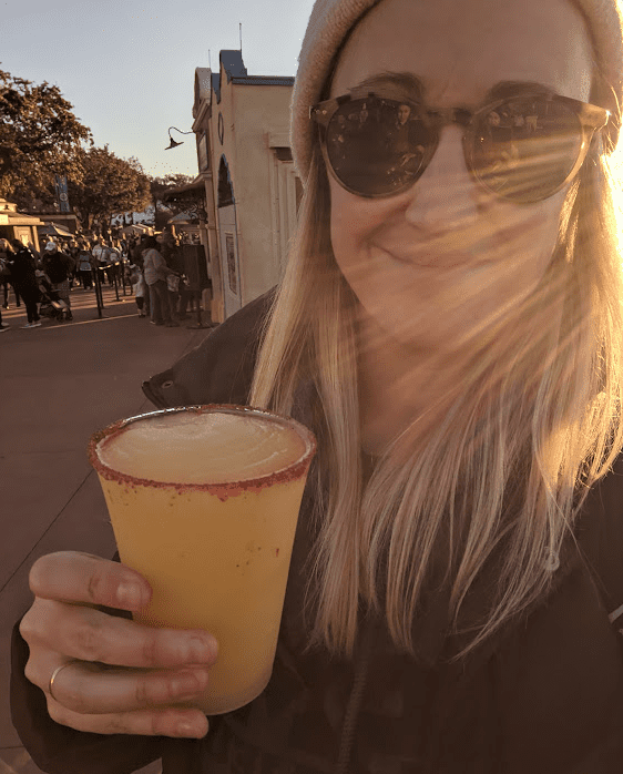 Girl in sunglasses and a beanie holding a frozen margarita and smiling at the camera. There’s an artistic lens flare partially over her face.