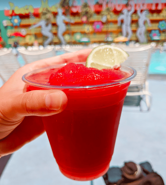 Hand holding a strawberry frozen margarita with a bright sky and sunny pool in the background. 
