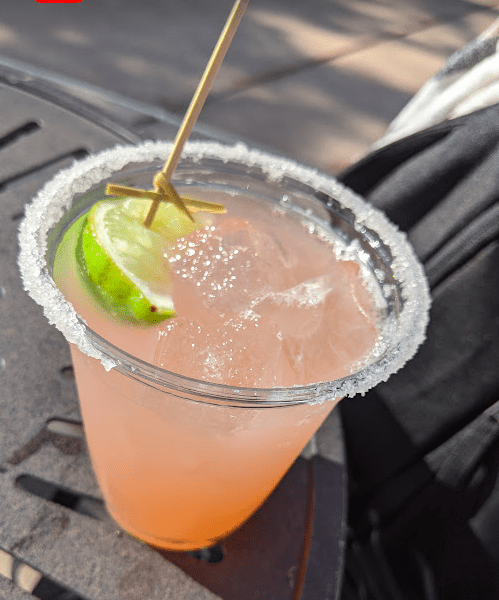 Pink margarita and a lime in a cup with a sugar-coated rim sitting on a metal table. 
