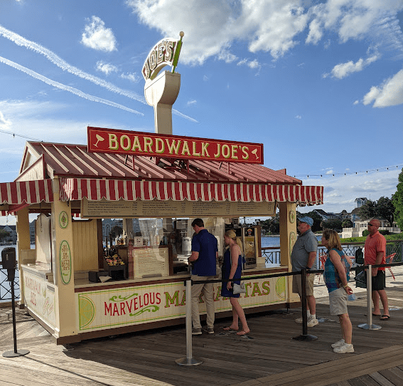 Boardwalk Joe’s margarita kiosk with “marvelous margaritas” painted on the side. Blue sky in the background and a small line of people in the foreground. 