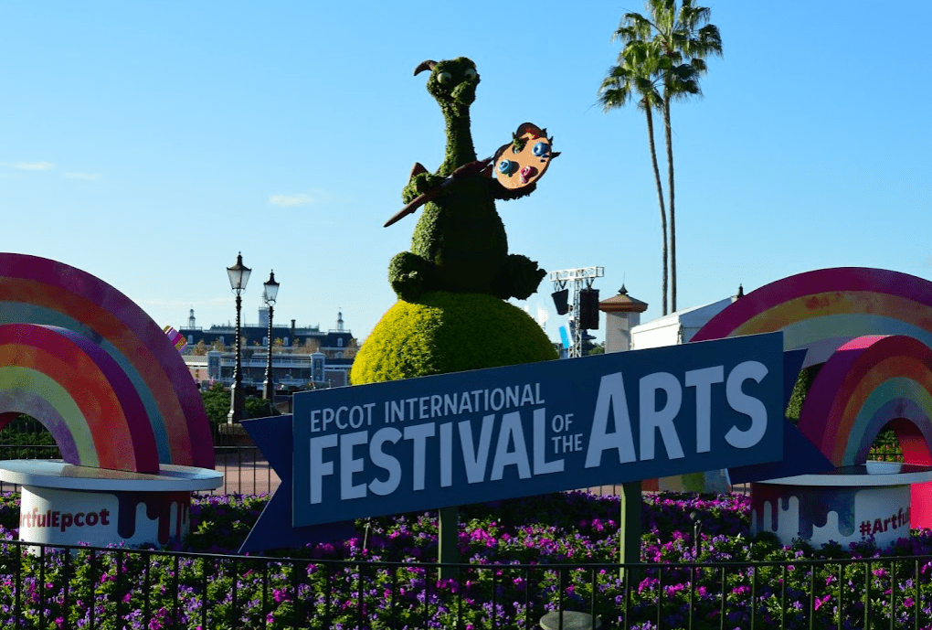 Figment topiary holding a paintbrush and palette sitting behind a sign that reads “EPCOT International Festival of the Arts.”
