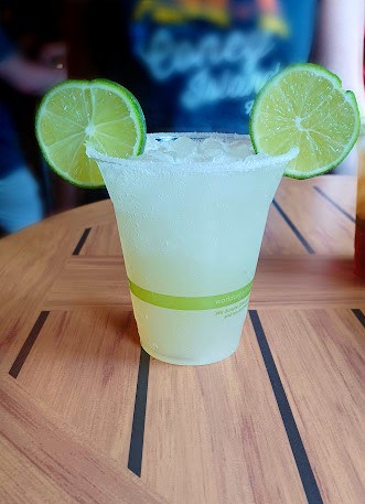 Classic margarita in a plastic cup with a salt rim sitting on a wooden table. The limes are positioned on either side of the rim to resemble Mickey ears.