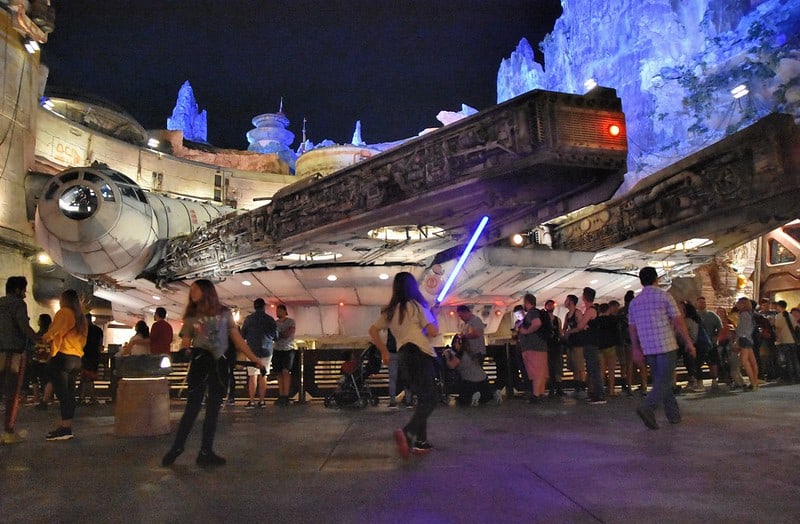 Millennium Falcon at Disney’s Galaxy’s Edge lit up at night with blue up-lighting on the rock formations around it. A child is mid-run in the foreground holding an illuminated light saber.