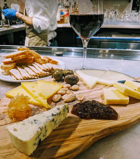 Loaded wooden cheese board sitting on the bar with a plate of bread/crackers and a glass of red wine at Wine Bar George in Disney Springs.