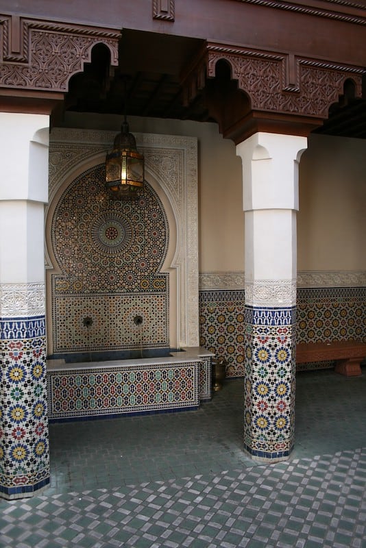Photo of an empty bench next to a colorful mosaic-tiled fountain and tiled pillars taken in the Morocco Pavilion at EPCOT. 