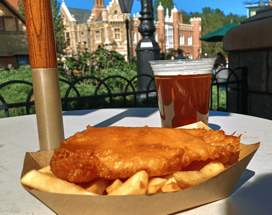 Fried fish on top of steak fries in a paper tray with an ale on a table. The United Kingdom Pavilion at EPCOT is in the background.